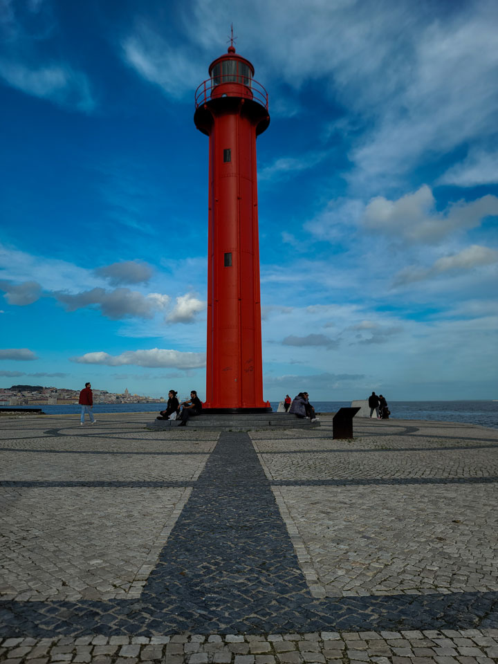 A striking red tower with a weathered exterior, covered in graffiti, stands along Rua do Ginjal in Cacilhas, Almada, Portugal. The abandoned structure, surrounded by urban decay and street art, contrasts beautifully against the backdrop of the Tagus River and Lisbon skyline.