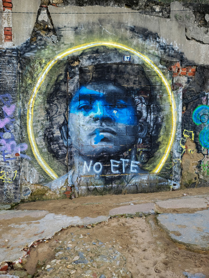 A vibrant graffiti mural of Diego Maradona on a weathered wall in Rua do Ginjal, Cacilhas, Almada, Portugal. The artwork, surrounded by colorful urban street art, portrays the legendary footballer in an expressive style, reflecting his passion and impact on the sport.